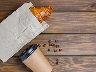 Coffee paper eco cup, beans and croissant in paper packaging on wooden background on the table. Perfect breakfast in the morning. Rustic style, top view