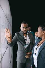 Black african businessman looking at female colleague pointing to large video screen for a...