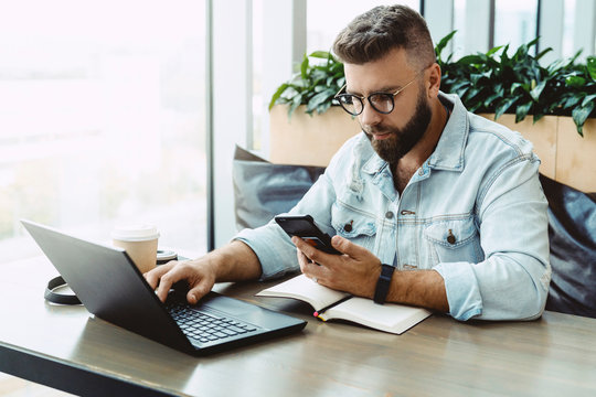 Hipster man sits in cafe, uses smartphone, works on laptop. Businessman reads an information message in phone. Freelancer works outside office. Teleworking. Online marketing, education for adult.