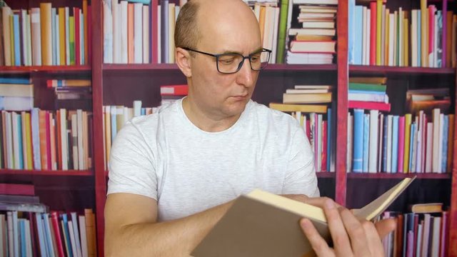 Thoughtful white man read book at bookshelf background in library interior. Man in glasses and white t-shirt sits at desk table and thinking with book. Close up portrait serious male face in glasses