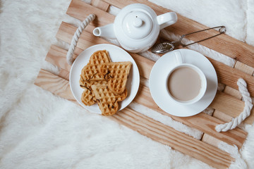 Cozy breakfast in bed, cup of coffee and heart shaped waffles on wooden tray on white and gray cozy blanket, the concept of home comfort, copy space