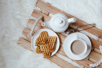 Fototapeta na wymiar Cozy breakfast in bed, cup of coffee and heart shaped waffles on wooden tray on white and gray cozy blanket, the concept of home comfort, copy space
