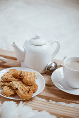 Cozy breakfast in bed, cup of coffee and heart shaped waffles on wooden tray on white and gray cozy blanket, the concept of home comfort, copy space