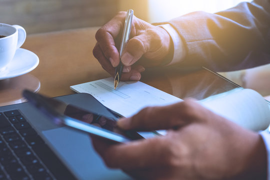 Man hand holding mobile smart phone and signing checkbook on the desk at office. Paycheck concept.