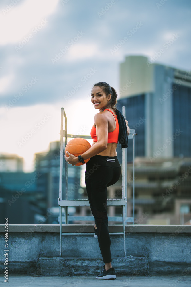 Wall mural Fitness woman standing on rooftop holding a basketball - Wall murals