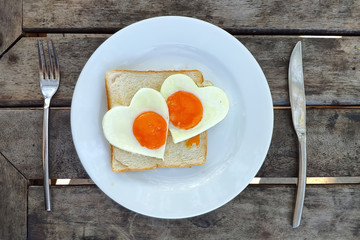 Breakfast for Valentines day. Fried eggs in the shape of a heart at toast on white plate and wooden table.  Knife and fork lie nearby