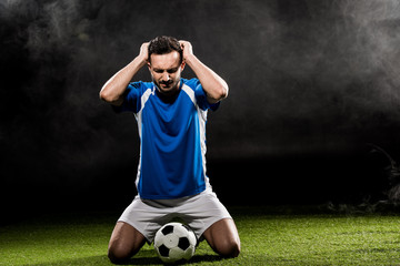 handsome football player holding head while sitting on grass on black with smoke