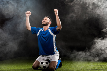 handsome football player celebrating victory while sitting on grass on black with smoke