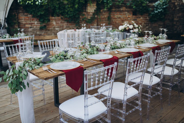 in the wedding banquet area there are tables and transparent chairs, on the tables there are plates, glasses, cutlery and compositions from green branches and candles