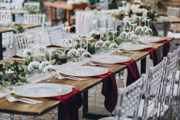 in the wedding banquet area there are tables and transparent chairs, on the tables there are plates, glasses, cutlery and compositions from green branches and candles