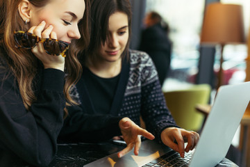 Fototapeta premium Two girls, friends Drink Hot Coffee While Work In Cafe On Laptop. Portrait Of Stylish Smiling Girls In Winter Clothes Drinking Hot Coffee And Work At Laptop. Female Winter Style. - Image