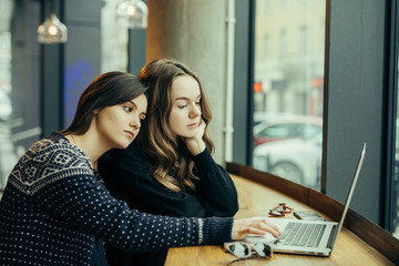 Two girls, friends Drink Hot Coffee While Work In Cafe On Laptop. Portrait Of Stylish Smiling Girls In Winter Clothes Drinking Hot Coffee And Work At Laptop. Female Winter Style. - Image