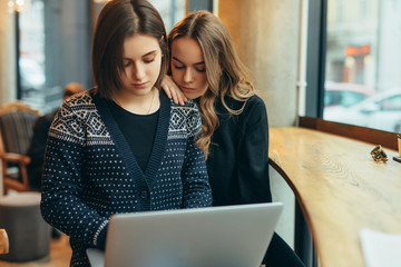 Two girls, friends Drink Hot Coffee While Work In Cafe On Laptop. Portrait Of Stylish Smiling Girls In Winter Clothes Drinking Hot Coffee And Work At Laptop. Female Winter Style. - Image