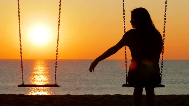 Back view silhouette of a sad girl at sunset missing her partner on a swing on the beach