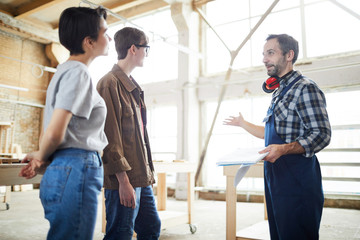 Fototapeta premium Confident bearded builder with ear protectors on neck gesturing hand and holding sketchpad while presenting house project to customers at construction site