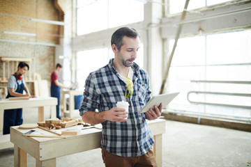 Busy pensive young joiner with safety goggles on tshirt leaning on table with wooden details and tools and drinking coffee while reading article on tablet