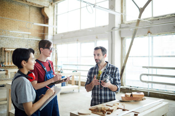 Content skilled handsome carpenter in checkered shirt standing at table with wooden pieces and showing power hand drill while teaching carpentry students in workshop