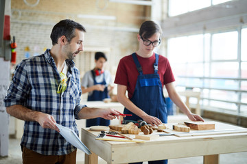 Serious handsome carpenter with stubble pointing at wooden piece and giving advice to student in...