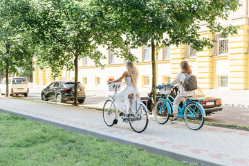 Fototapeta premium Two blonde friends ride on bicycles along the boulevard/ white and blue retro bikes with baskets, sensuality and elegance, healthy and happy cyclist women/ summertime, leisure and lifestyle concept.