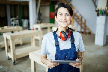 Smiling attractive young female carpenter with ear protectors on neck standing in carpentry...