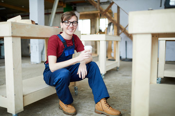 Positive handsome young carpenter in classes sitting on wooden part of wheeled table and looking at camera while drinking coffee during break