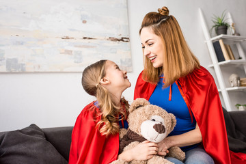 smiling mother and kid in red cloaks looking at each other and holding teddy bear
