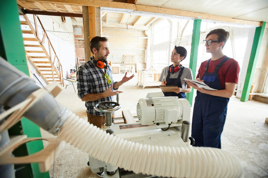 Serious confident handsome carpenter with ear protectors on neck standing in workshop and talking to interns while explaining then how to work with modern machine