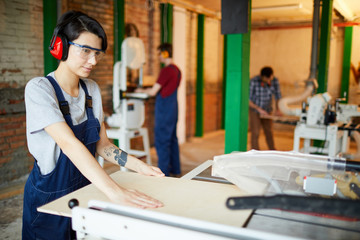 Serious concentrated young lady in safety goggles and ear protectors standing at workbench and adjusting wooden plank to cut it on saw table