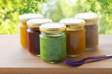 baby food, healthy eating and nutrition concept - vegetable or fruit puree in glass jars and feeding spoon on wooden table over green natural background