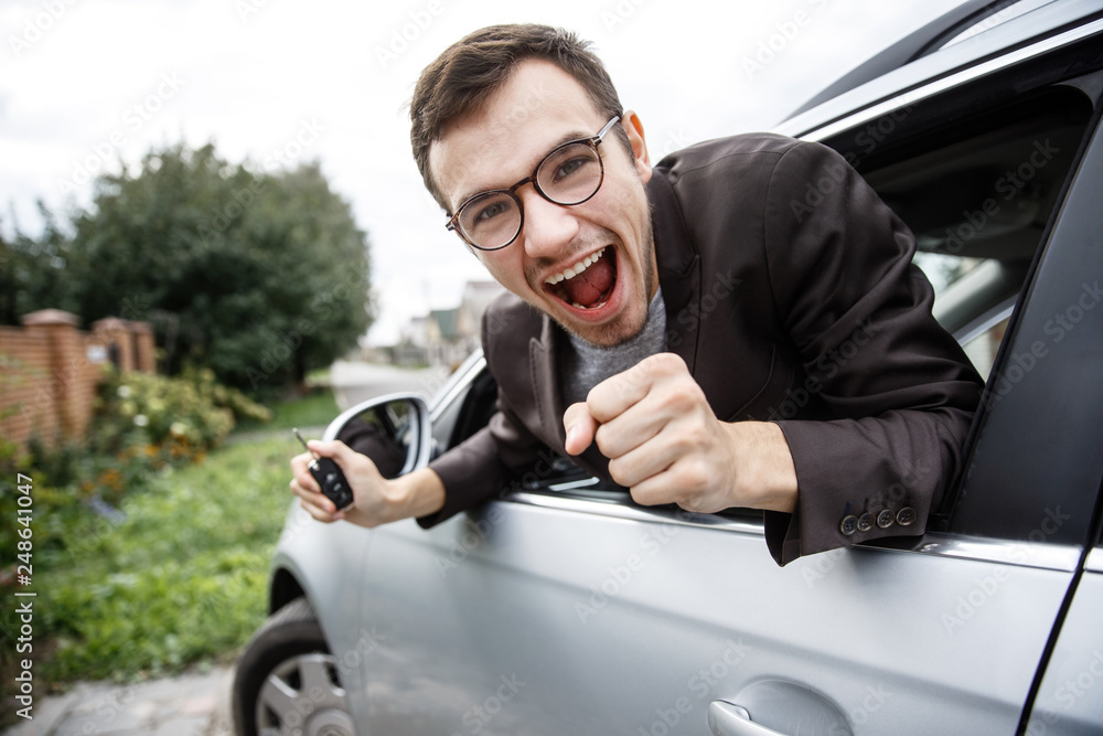 Wall mural obsessed young guy is peeking from the car window while looking at the camera. he is holding the key