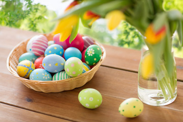 Fototapeta na wymiar easter, holidays, tradition and object concept - colored eggs in basket and tulip flowers on wooden table over green natural background