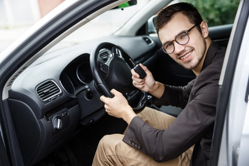 Satisfied young driver is sitting at his car while looking at the camera. He is holding the keys at his right hand. His left hand is on the steering wheel.