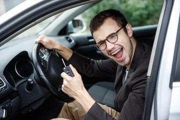 Very happy young man is sitting at his car while looking at the camera. He is holding the keys at his left hand. His right hand is on the steering wheel.
