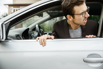 Angry young man is peeking from the window. He is looking at something behind his car.