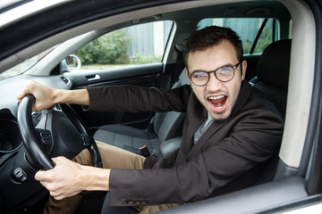 Fototapeta na wymiar Furious young man is looking at the camera while sitting at his car. He is screaming at someone. His hands are on the steering wheel. Angry driver concept.