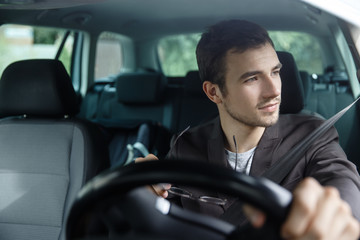 Delighted young man is looking trough the window while driving his car. He is holding his glasses at his right hand. His left hand is on the steering wheel.