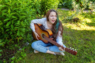Young hipster woman sitting in grass and playing guitar on park or garden background. Teen girl...