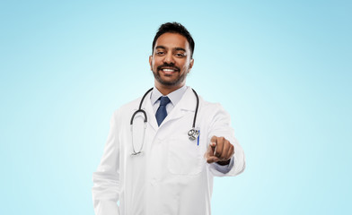medicine, profession and healthcare concept - smiling indian male doctor in white coat with stethoscope pointing finger to you over blue background