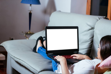 Young woman lying comfortably on the sofa while using the laptop with blank screen. View from behind.