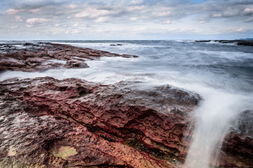 Sea waves hit the red rocks of the beach with clouds and sky in the background. 