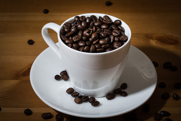 White cup filled with coffee beans  and coffee beans scattered on a wooden surface and on the white saucer. 