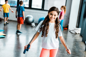 Cute brunette kid holding jump rope and smiling at camera - obrazy, fototapety, plakaty