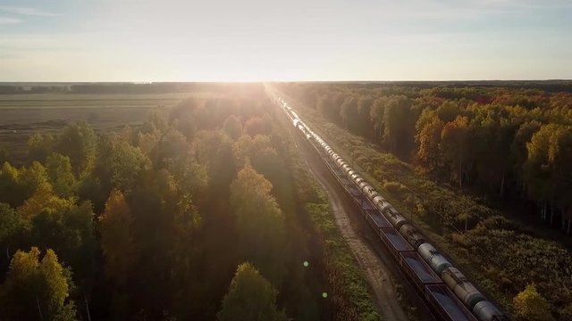 upper view freight trains with cisterns and open wagons move in opposite directions through forest against bright sunset