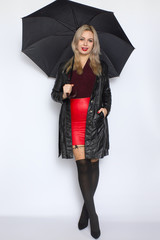 Full length portrait of blonde woman in leather black coat and red skirt holding open black umbrella in her hands. Over white background in studio