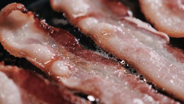 Crispy, roasted bacon slices on a frying pan. Camera panning over a close-up view of cooked, crispy, roasted bacon slices with bubbly cooking oil on a stove.