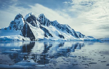 Gordijnen Met ijs bedekte bergen in de polaire oceaan. Winter Antarctisch landschap in blauwe en witte tinten. De weerspiegeling van de berg in het kristalheldere water. De bewolkte hemel over de enorme gletsjer. Reis door de wilde natuur © Goinyk