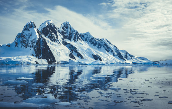 Ice covered mountains in polar ocean. Winter Antarctic landscape in blue and white tints. The mount's reflection in the crystal clear water. The cloudy sky over the massive glacier. Travel wild nature