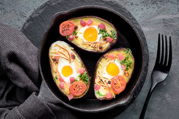 Keto diet dish: avocado boats with ham cubes, quail eggs, cheese and cherry tomatoes on iron cast skillet