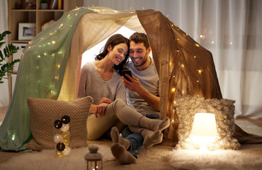 Obraz na płótnie Canvas leisure, hygge, technology and people concept - happy couple with smartphone in kids tent at home