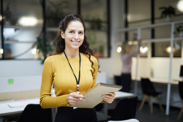 Smiling pretty young lady with badge on neck standing in modern open space office and holding...
